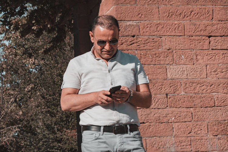 Man using smart phone standing against brick wall