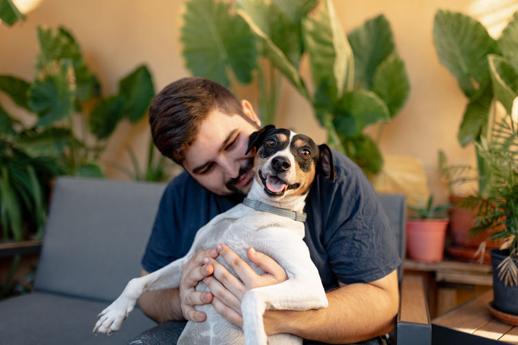 Young man smiling is holding up his dog while playing with his paws. the dog is looking at camera
