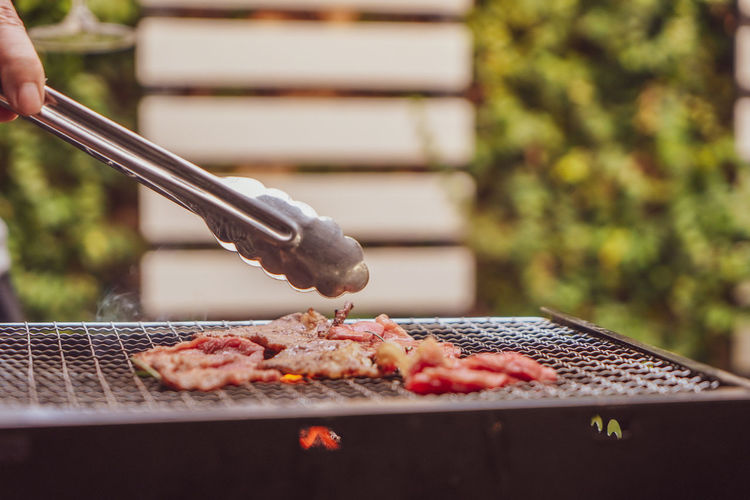Cropped hand using meat tongs to grill slices of premium meat on the stovetop at 
 outdoor party. 