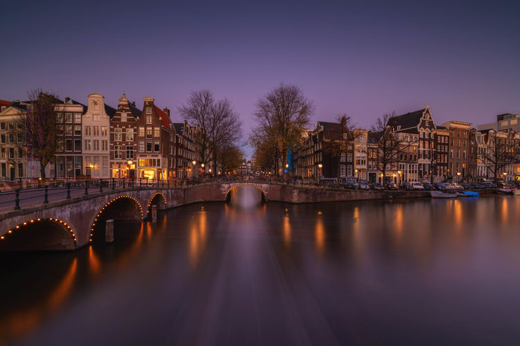 Bridge over river amidst illuminated buildings in city at dusk