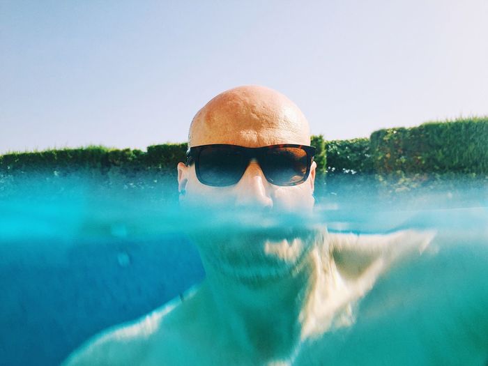 Portrait of bald man wearing sunglasses while swimming in pool against clear sky