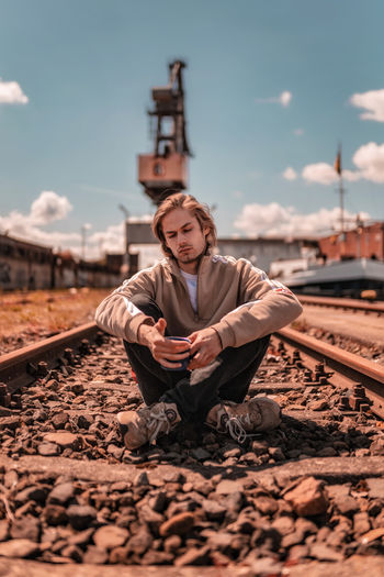 Young man sitting on railroad track against sky