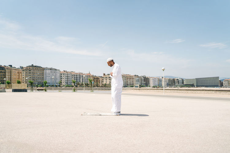 Full boy islamic male in traditional white clothes standing on rug and praying against blue sky on beach