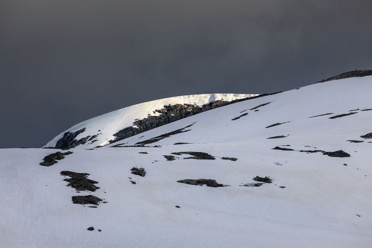 Snowcapped top of a mountain spot lit by sun against dark grey moody sky