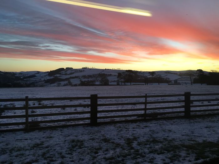 Fence on snow covered field against cloudy sky during sunset