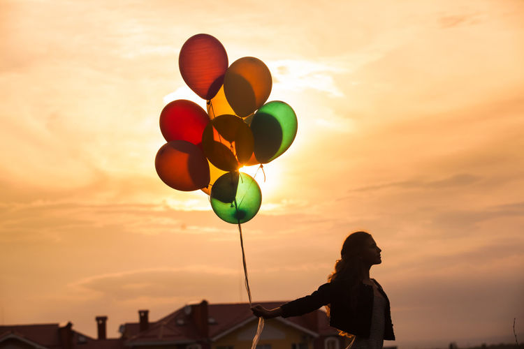 Silhouette of person holding balloons against sky during sunset