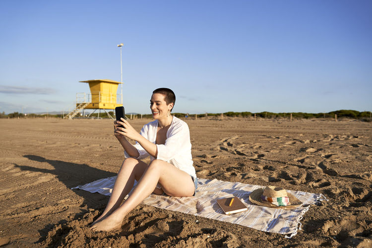 Smiling woman using mobile phone while sitting at beach