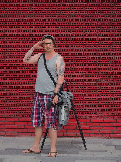 Hipster with camera saluting while standing on sidewalk against red wall