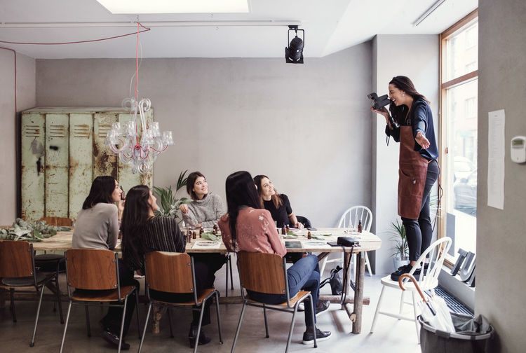 Cheerful woman standing on chair photographing female colleagues sitting at table in workshop