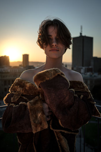 Portrait of young woman standing by railing in city against sky during sunset