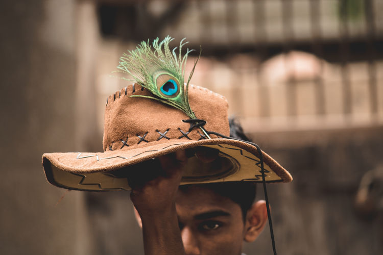 Close-up of teenage boy holding hat and peacock feather while standing against wall