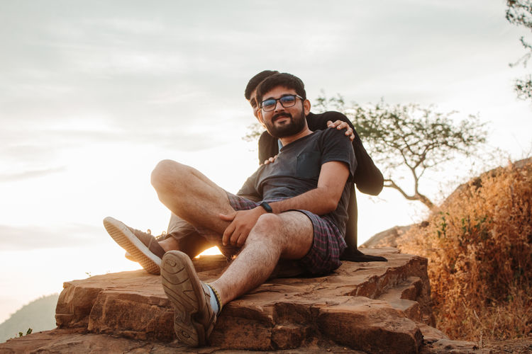 Portrait of young man with friend sitting on rock against sky