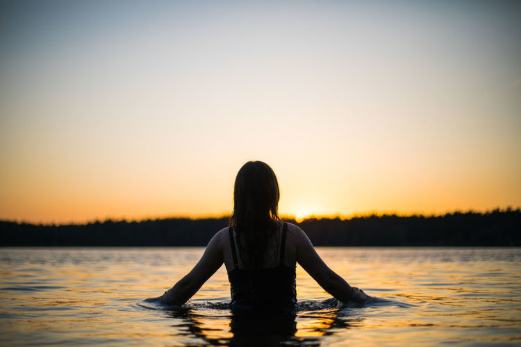 Girl in a long black swimsuit swims outside the city on the lake in the rays of sunset or dawn. 