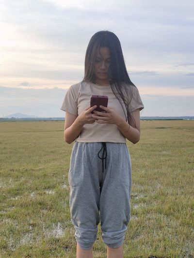 Young woman using mobile phone while sitting on field against sky