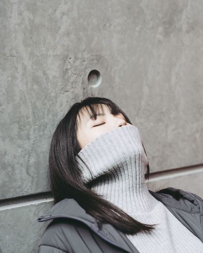 Close-up of young woman covering mouth with turtleneck against wall