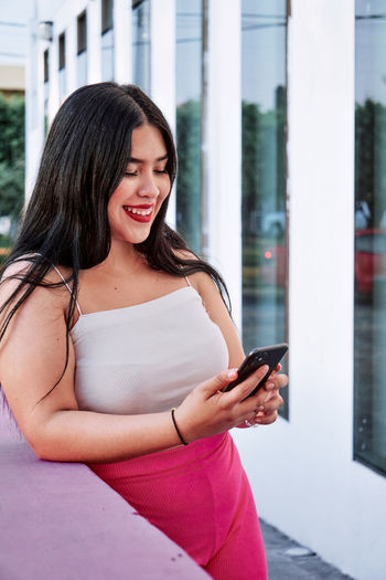 Happy overweight ethnic woman with long black hair and red lips smiling while leaning on border near modern building and browsing phone on city street