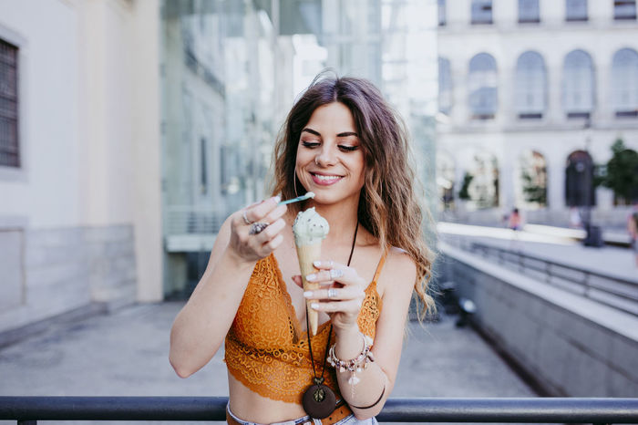 Smiling woman having ice cream while standing against building in city