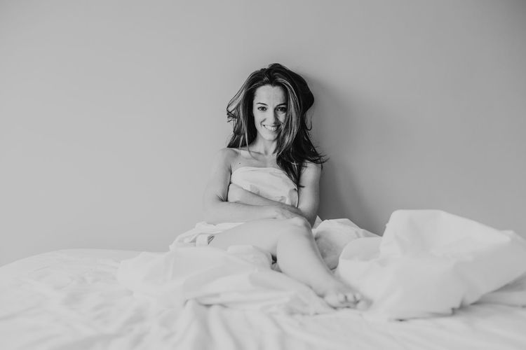 Portrait of smiling woman with blanket sitting on bed against wall