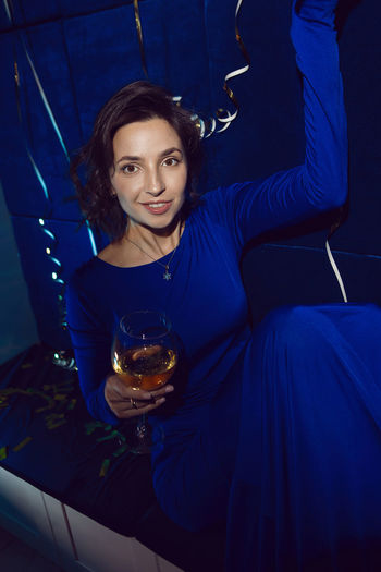 Cheerful caucasian brunette girl in a blue dress with a glass of champagne, sitting on a sofa