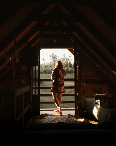 Rear view of woman on ledge of wooden cabin at sunset
