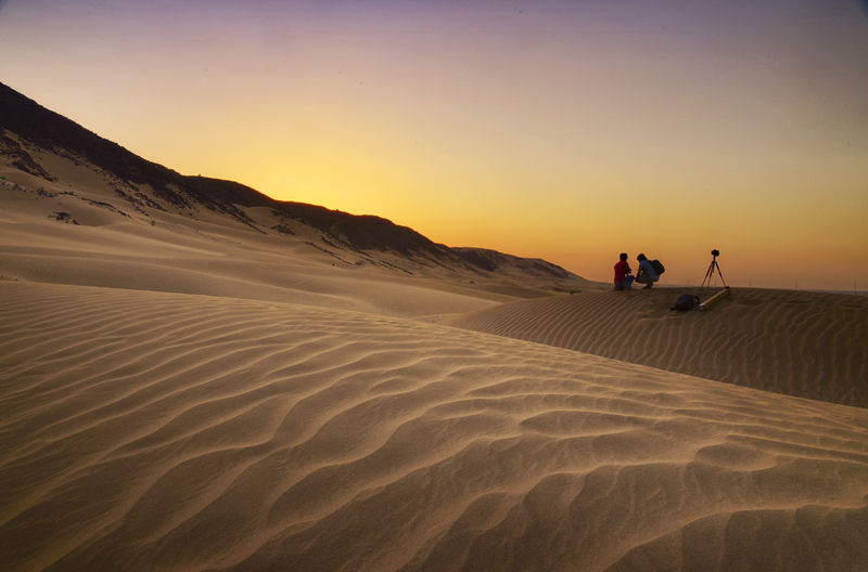 People on sand dune in desert against sky during sunset