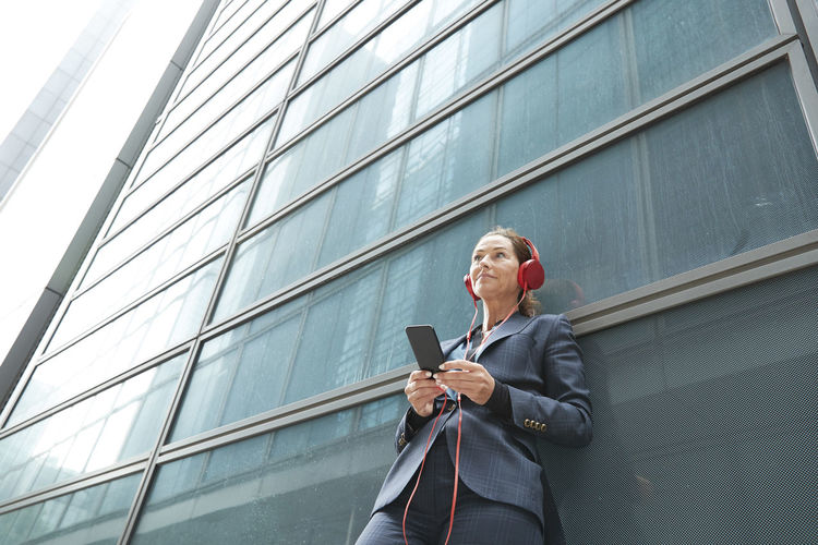 Businesswoman with headphones listening music while standing against building in city