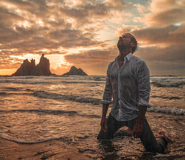 Man kneeling on beach against sky during sunset
