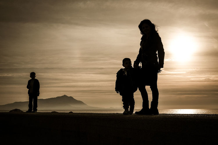 Silhouette mother with sons standing at beach against sky during sunset
