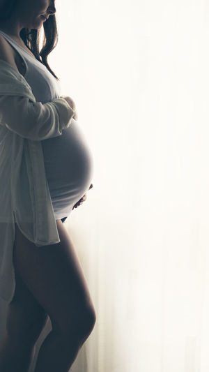 Midsection of pregnant woman standing by curtain at home