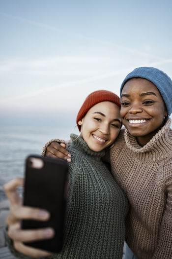 Young women wearing knit hat taking selfie on mobile phone while standing against sky