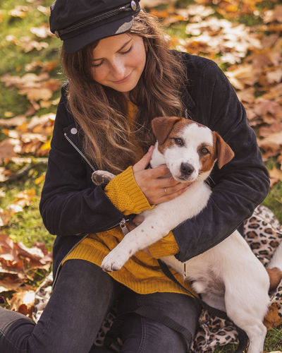 Smiling woman holding dog sitting at park during autumn