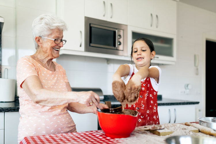 Girl with dough on hands against cheerful grandma at table with bowl during cooking process at home