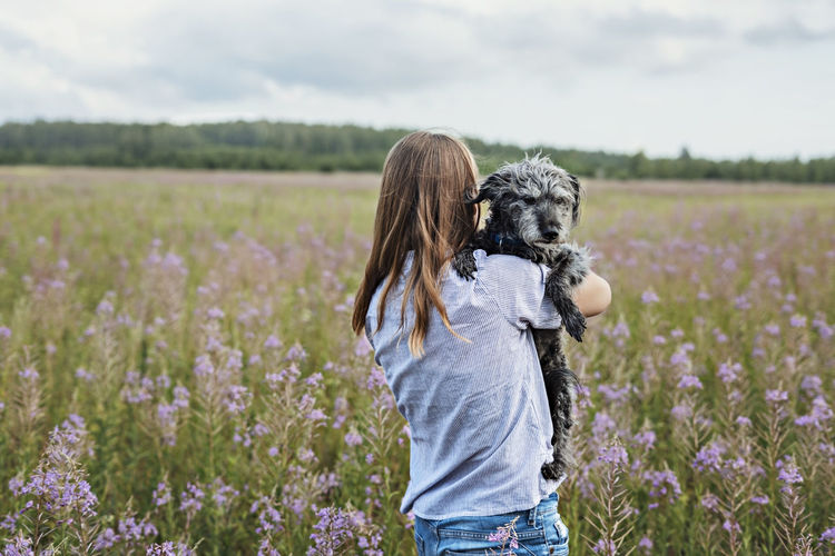 Rear view of young woman holding fluffy senior dog in hands on field of flowers fireweed , pet love
