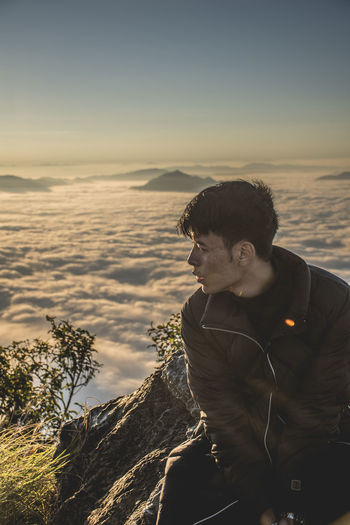 Young man looking away while sitting on mountain against clear sky during sunset