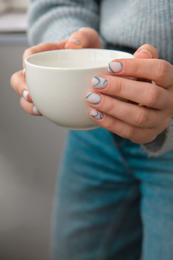 Manicured female hands holding white coffee cup with stylish blue nails and minimalistic design. 