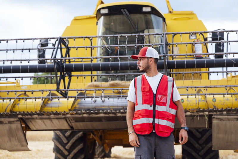 Bearded male farmer in uniform looking away while standing on agricultural field with industrial combine harvester during work in countryside