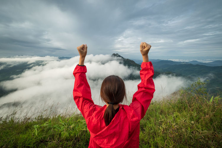 Rear view of woman with arms raised looking at landscape against cloudy sky