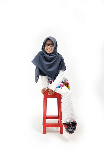 Portrait of smiling young woman wearing hijab sitting on stool against white background