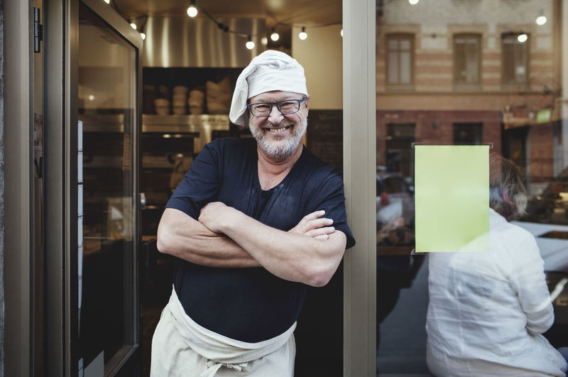 Smiling male baker standing with arms crossed at entrance of bakery