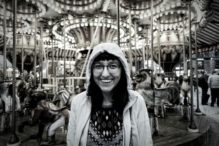 Portrait of smiling young woman against carousel horses at amusement park