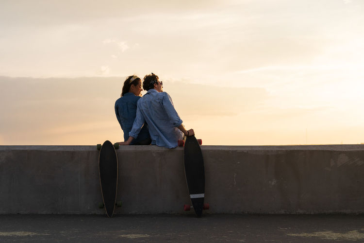 Stylish couple of longboarders at seaside sit on pavement watch summer sunset. lifestyle concept