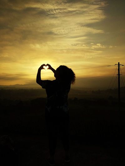 Silhouette woman making heart shape standing against sky during sunset