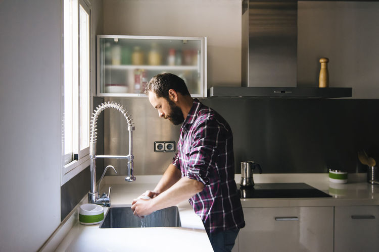 Man with beard and plaid shirt washing dishes at home.