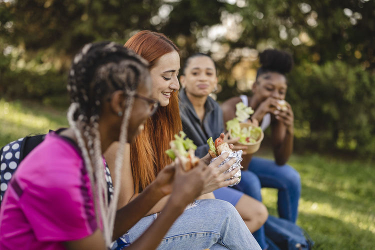 Smiling young woman with friends eating snack sitting in park
