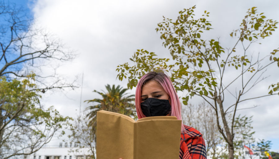 Low angle view of woman with mask reading book against trees