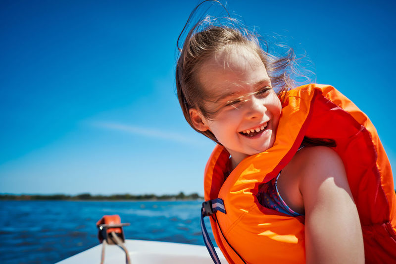 Close-up of cheerful girl wearing life jacket sitting against clear blue sky