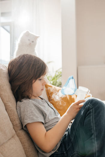 A little boy makes an inhalation at home sitting on the couch using a nebulizer and a mask. 