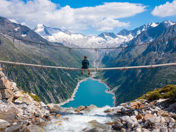 Man sitting on bridge over valley against snowcapped mountains against sky