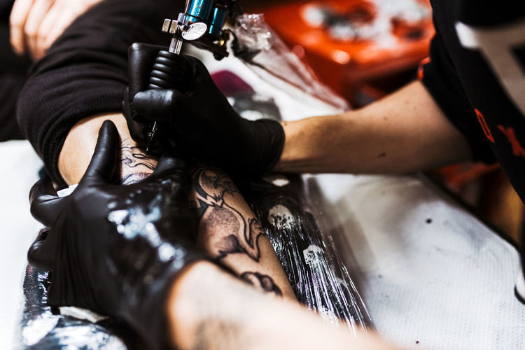 From above cropped unrecognizable man with using tattoo machine to make tattoo on leg of crop customer during work in salon