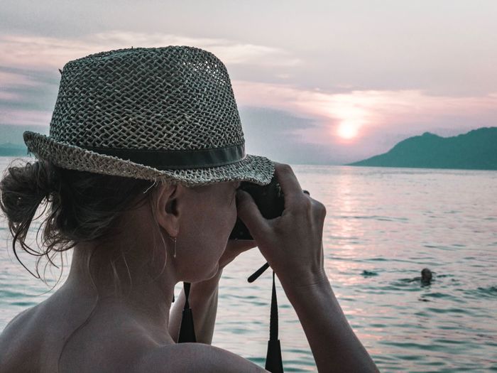 Close-up of woman wearing hat photographing sea with camera during sunset